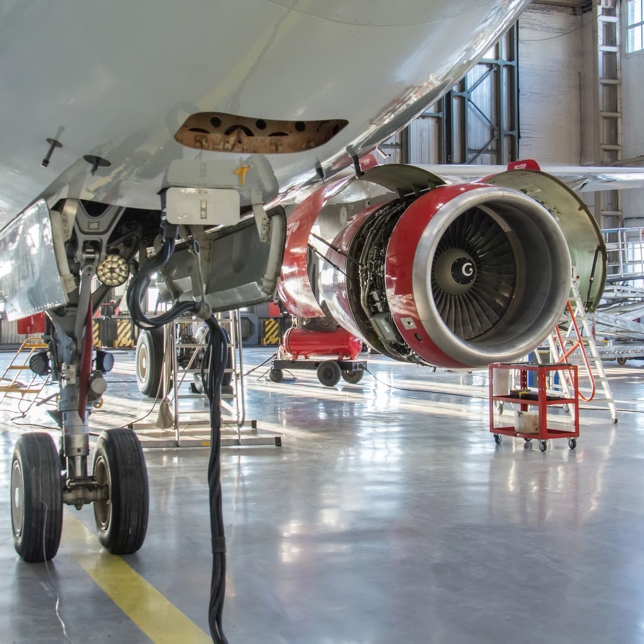 The front landing gear of an aircraft in service hangar in the background is an engine with an open hood